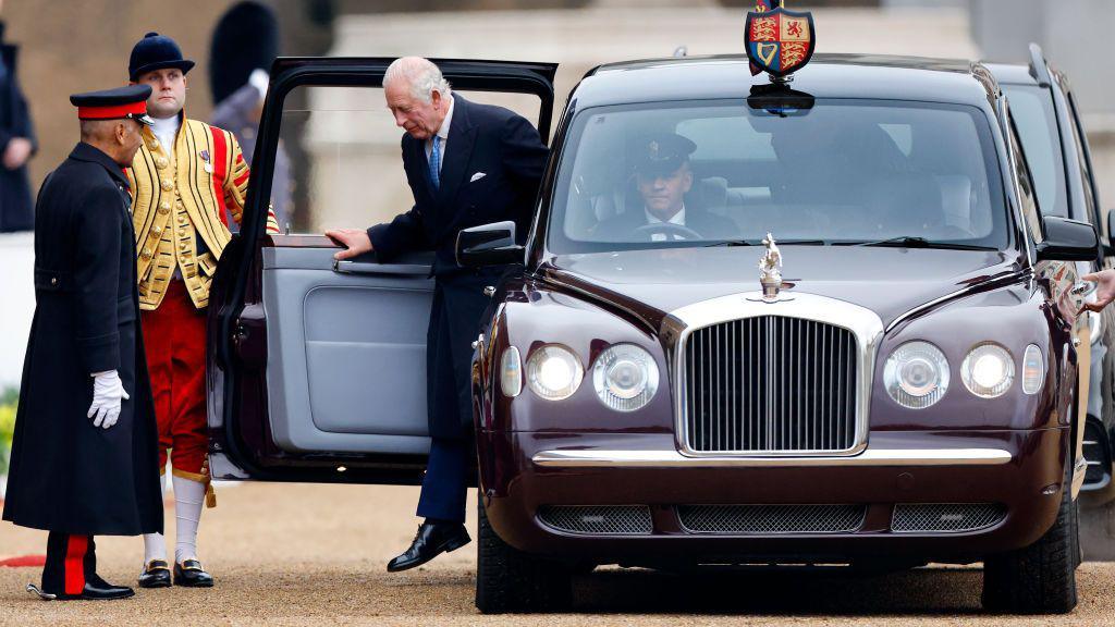 The King steps out of an official car which has a coat of arms on the roof of the vehicle. A liveried servant opens the door for him. 