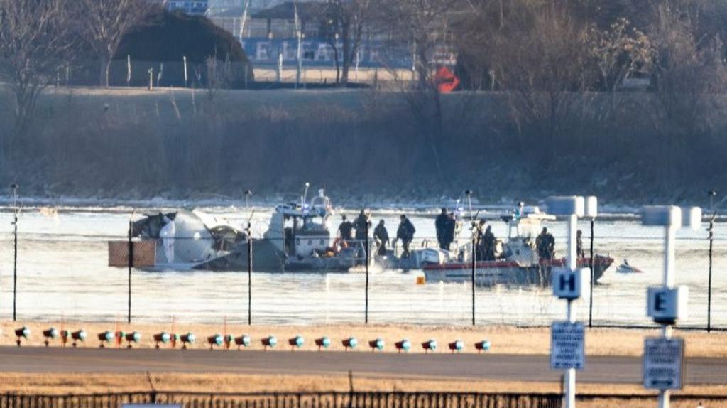 Two boats with people on them next to the wreckage of the plane in the river, seen from behind a fence and a runway at the airport