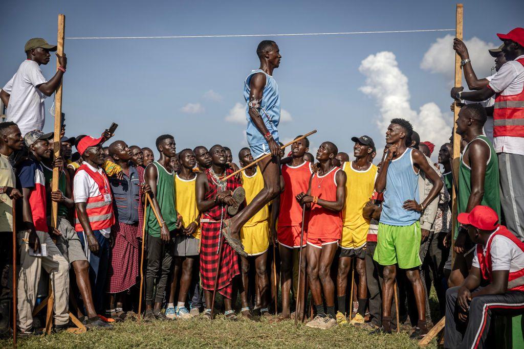 A Maasai athlete leaps vertically while taking part in the men's Maasai jumping competition, watched at close quarters by a number of spectators, some of whom are holding the high-jump frame.