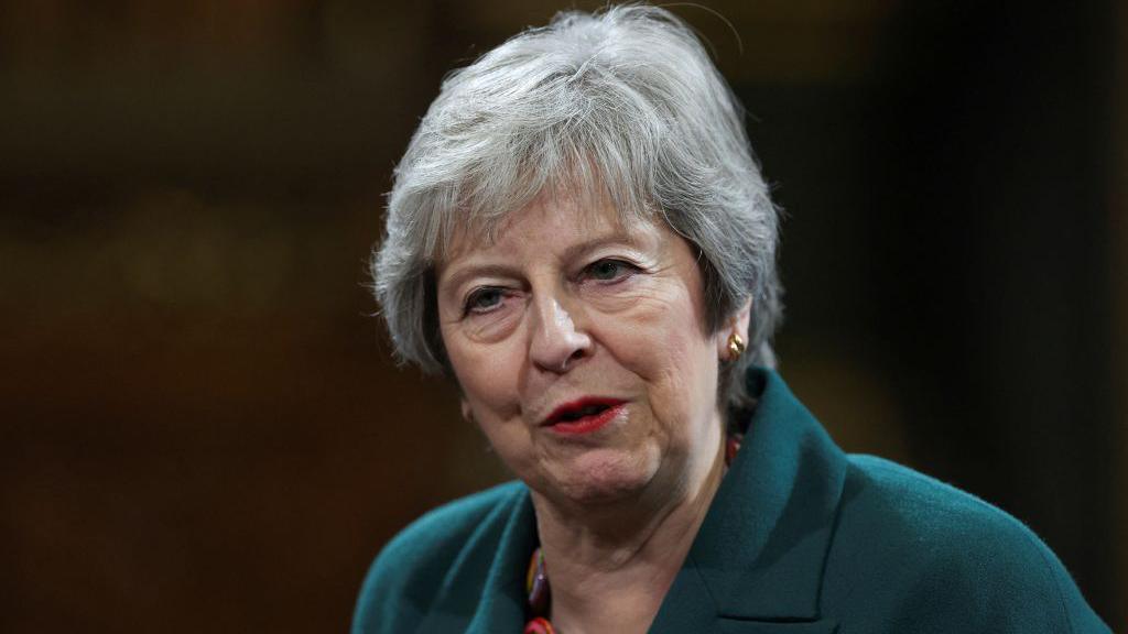 Theresa May with grey hair and wearing a forest-green blazer speaking to the camera