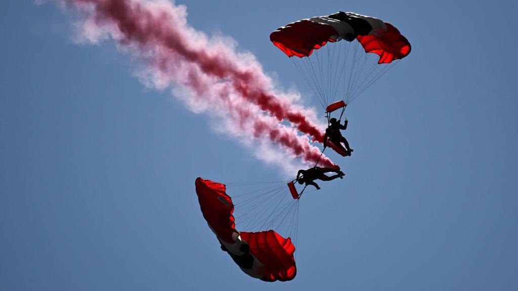 Two parachutists in the sky with red parachutes and red smoke coming off them.