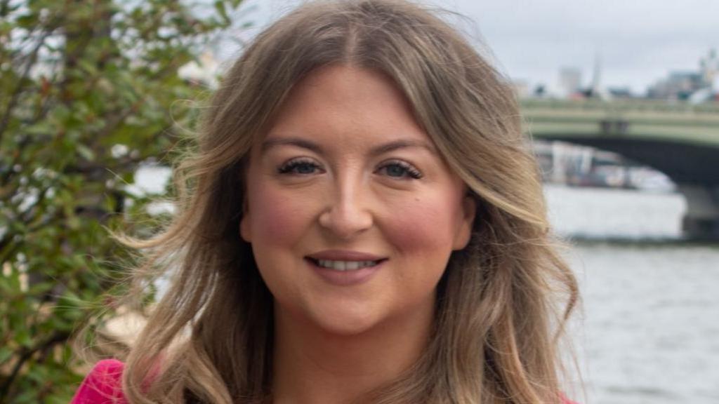 A woman with long blonde hair and wearing a cerise dress is smiling, with the River Thames in London in the background.
