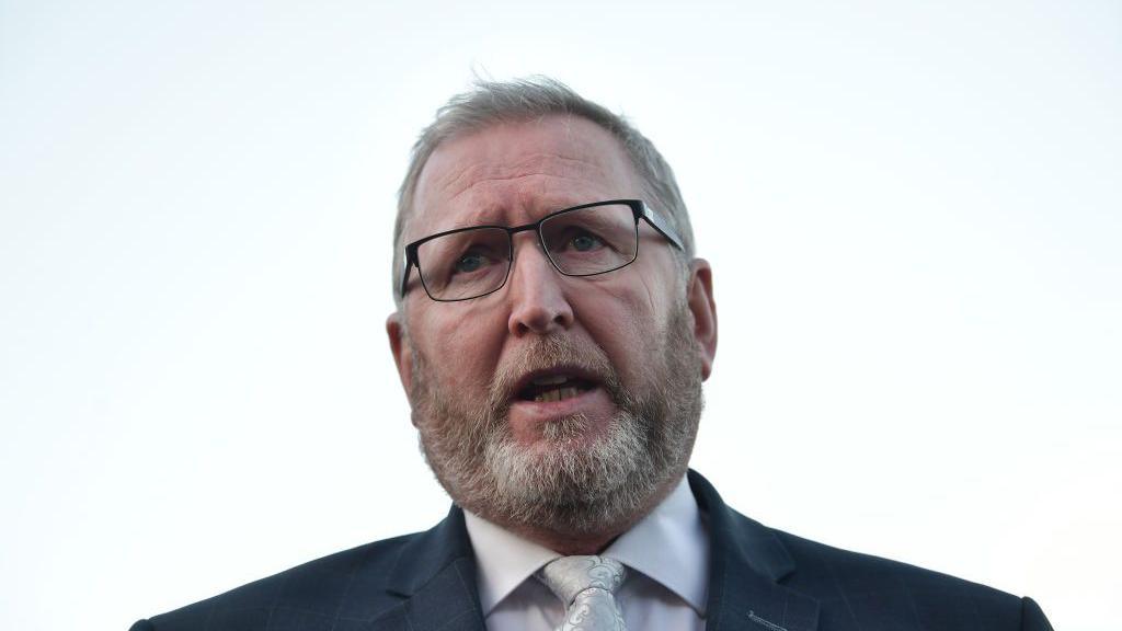 A man in a dark suit, white shirt and silver tie. He has short grey hair and a grey beard. He is wearing glasses, he is looking out over the top of the camera shot.