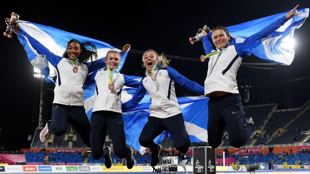 Four women, wearing tracksuits and holding Scottish flags, jump into the air while celebrating winning medals. They are in an athletics stadium.