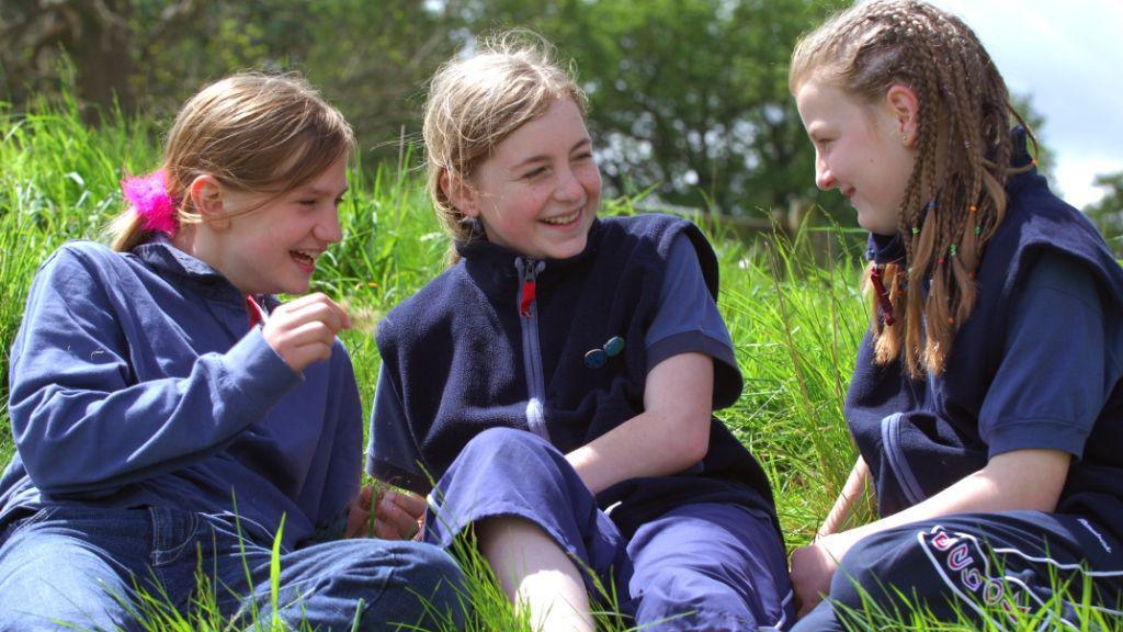 Photo shows three girl guides laughing together whilst sitting on the grass outside. They are wearing their navy blue uniforms with badges on, and one has a fluffy pink scrunchie in her hair.