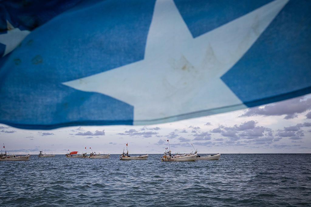 Somalia's national flag - bright blue with a white, five-pointed star in the middle - is blown by seawinds. The background boats are visible.