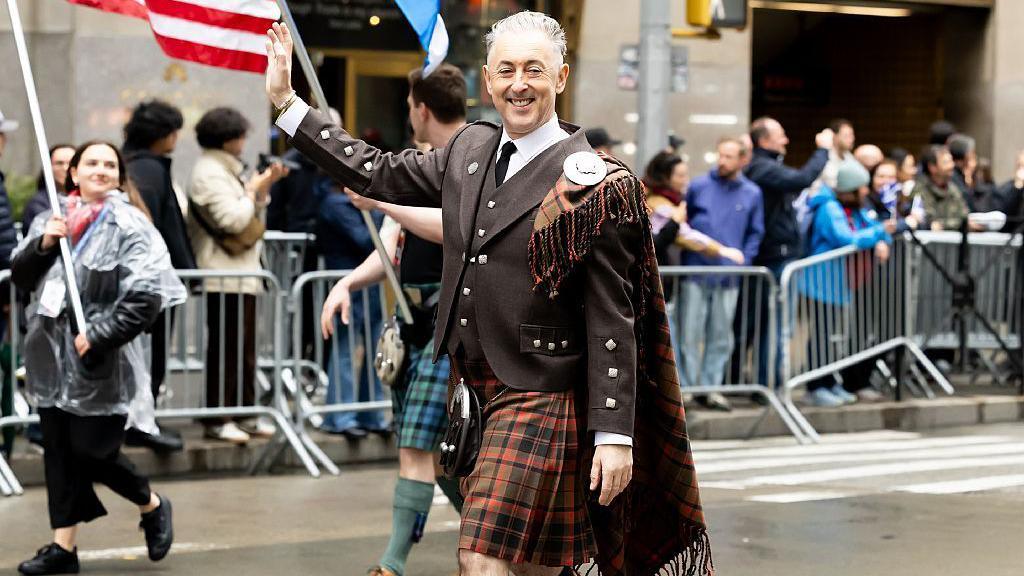 Alan Cumming, in brown and soft red Highland suit and kilt, is smiling and waving while walking along the middle of the street in New York. People are cheering in the background.