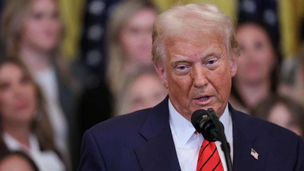 U.S. President Donald Trump speaks at an event in blue suit and red tie with blurred crowd behind him