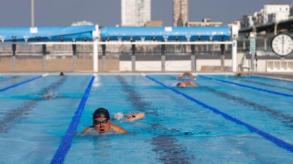 A man in black swimming hat and goggles swimming in an outdoor pool. 