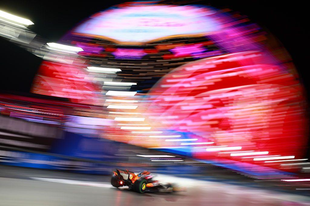 A Formula 1 car speeds through a brightly-lit Las Vegas night circuit, captured with dramatic motion blur against a huge, swirling neon display that fills the background with vibrant pinks, reds and purples.