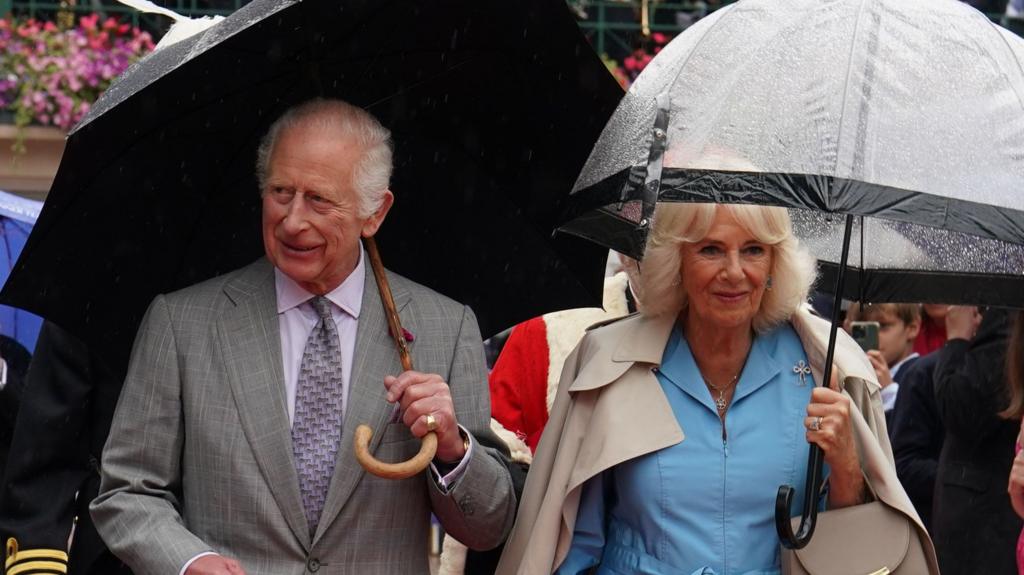 King Charles III and Queen Camilla carrying umbrellas and walking through Jersey's Royal Square