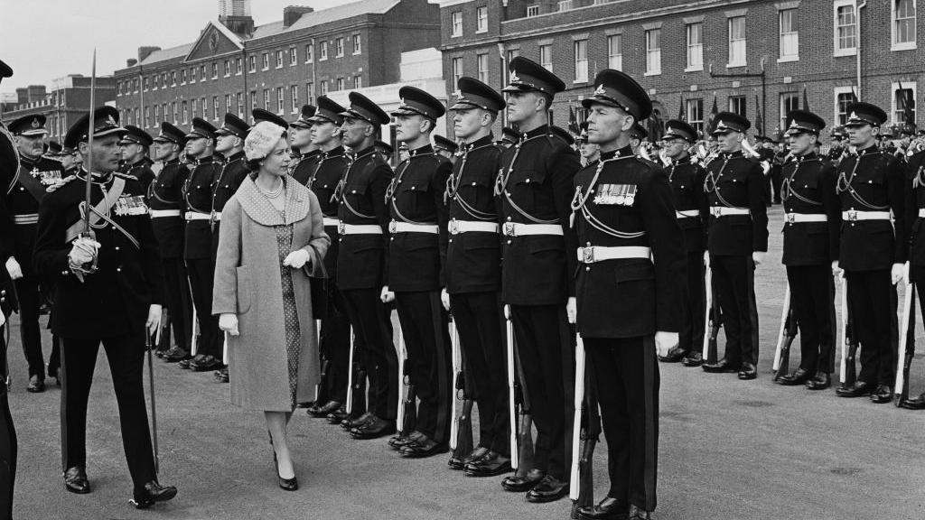 A black and white image of Queen Elizabeth II inspecting a row of British Army troops standing to attention outside Woolwich Barracks. 