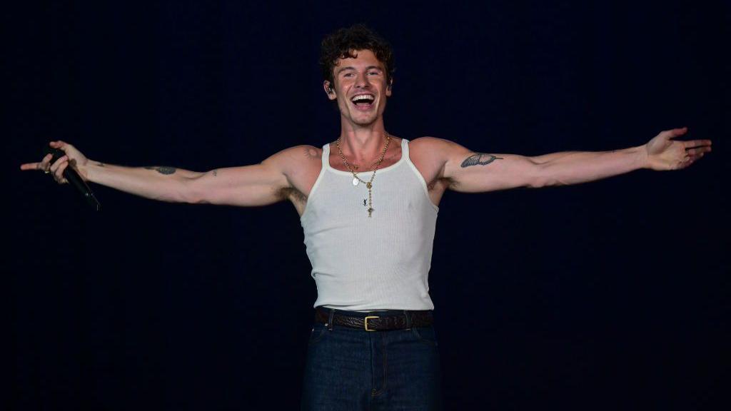 Shawn Mendes stands on stage smiling with his arms open wearing a white vest top and jeans.