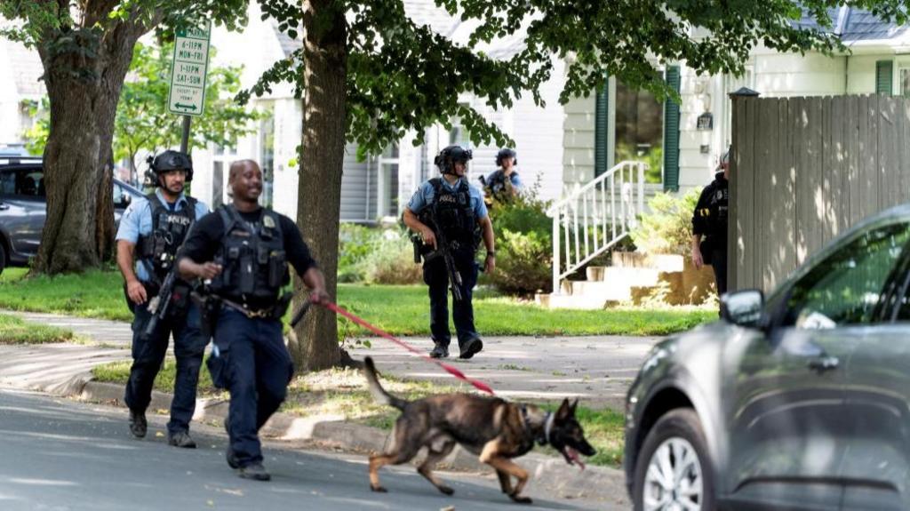 Heavily armed police searching through residential front gardens with a police dog.