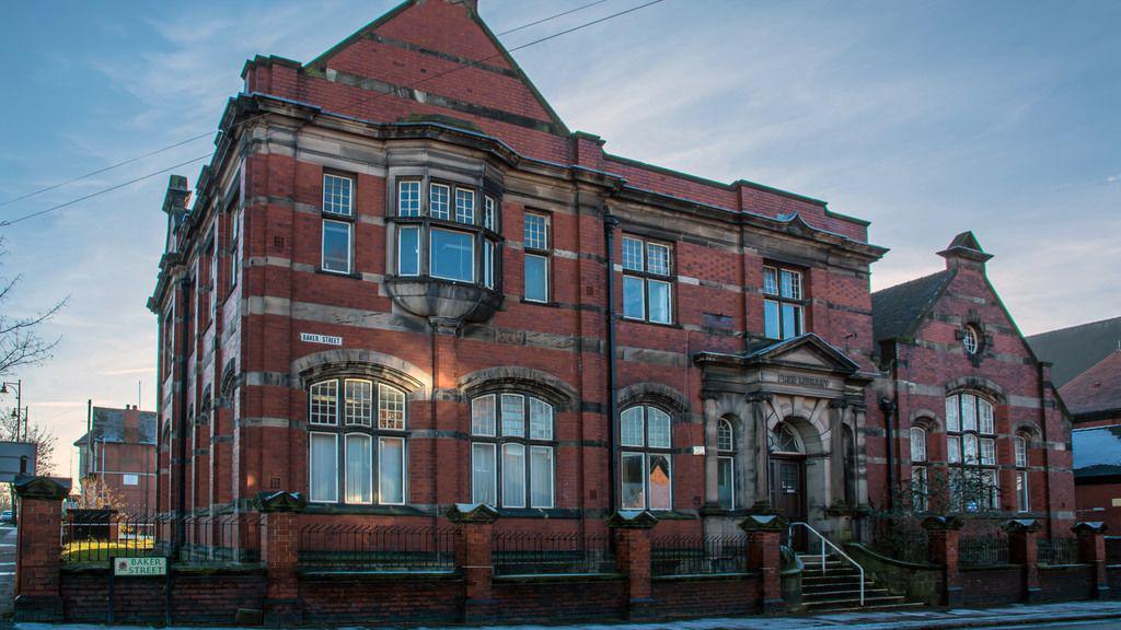 The front of an old two-storey library building. It is situated behind a fence and a wall, with steps leading up to an entrance.