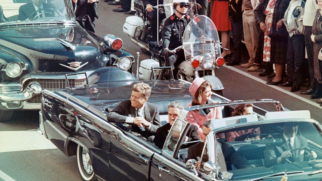 President Kennedy, pictured riding in an open convertible minutes before he was shot by a sniper. He is accompanied by his wife, who is wearing pink, and police officers. The are crowds of people lining the streets to see him