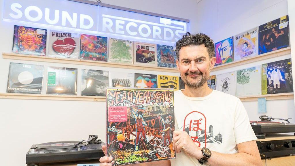 Tom Berry holds up a record with a colourful sleeve. He is wearing a white T-shirt and a black watch. Behind him various record players can be seen, with many records displayed on a white wall. The name of the shop, Sound Records, is written on a window near the ceiling in white block letters. 