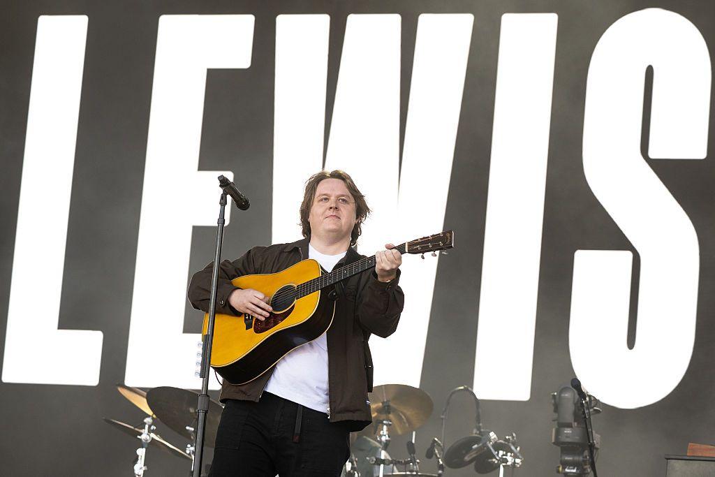 Lewis Capaldi, a man in his 20s with longish light brown hair sings on stage while playing an acoustic guitar. A screen lit up with his name "Lewis" fills the area behind him in black and white