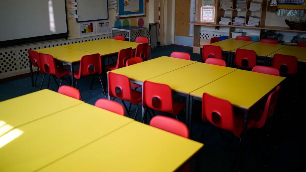 A stock image shows an empty classroom containing several yellow desks surrounded by red chairs.