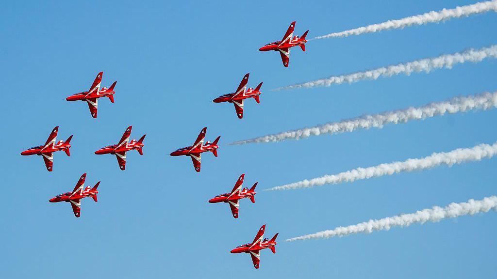 The famous Red Arrows jets - nine in total - fly from right to left across the picture in close formation with white smoke billowing from five of them at the back