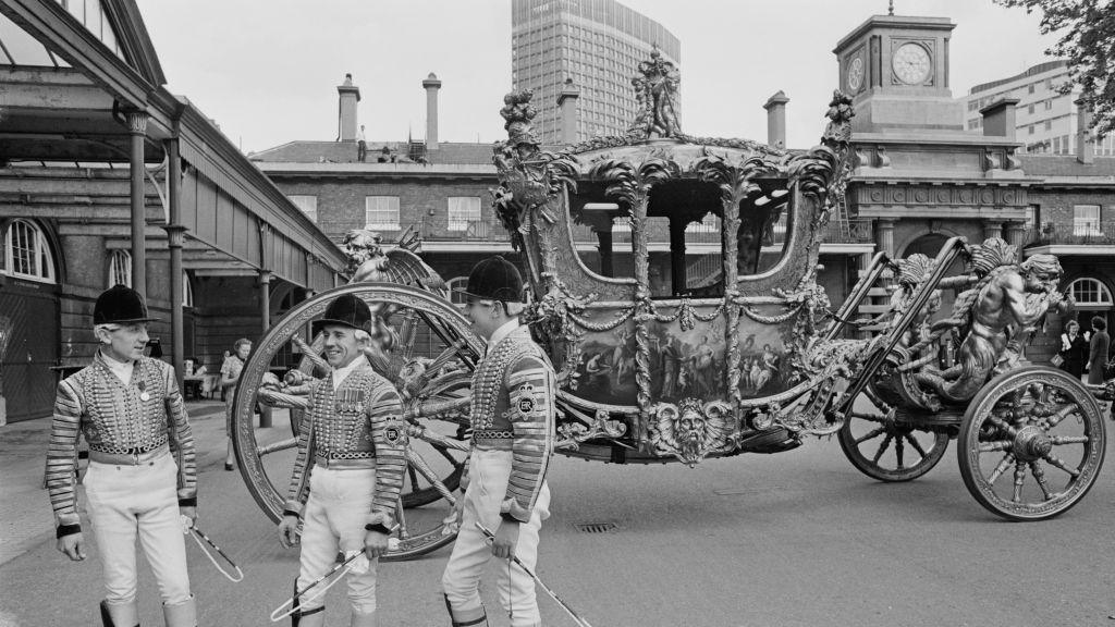 Three liveried coach drivers stand in the courtyard beside the Gold Coach. There is a modern tower block behind the Mews building. 