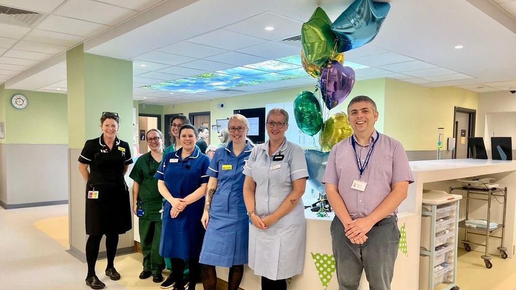 Medical professionals - all but one in uniform - lined up in front of a hospital reception area with balloons behind them. All are smiling at the camera.