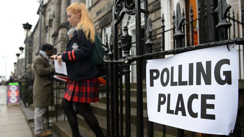 A young woman wearing a red and black tartan skirt leaves a polling station in Edinburgh, Scotland, during the 2014 independence referendum. A sign reading: Polling place is attached to black railings.