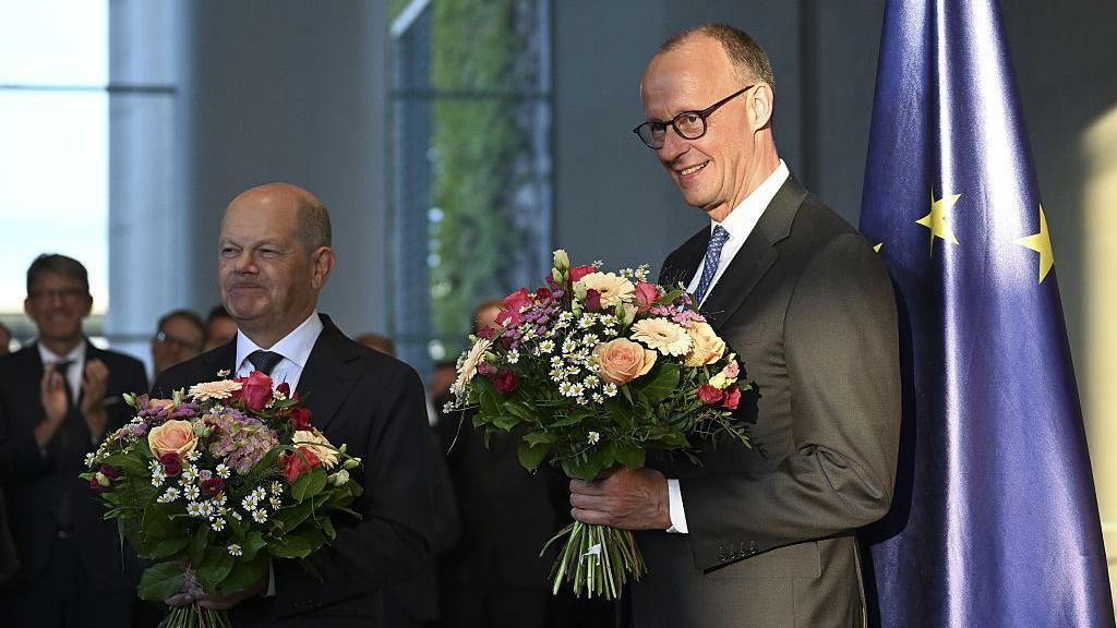 Olaf Scholz on left smiling and holding a bouquet of flowers. Friedrich Merz is on the right, also smiling and holding a bouquet. Behind him is a European Union flag.