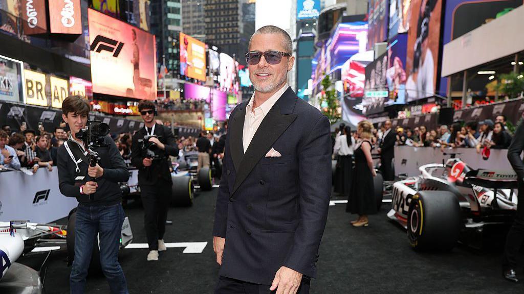 Brad Pitt stands in a black suit in front of multiple F1 racing cars in New York City at a promotional event for his film F1. Electonic billboards can be seen behind him on the streets of Times Square - including one for F1.