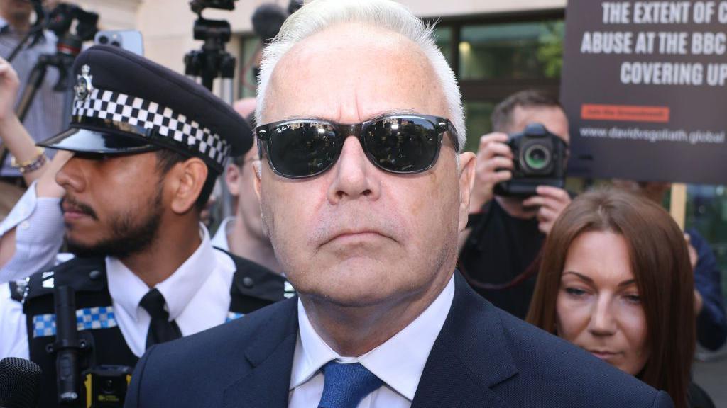 Huw Edwards in a suit and dark glasses leaves Westminster Magistrates' Court surround by reporters and police