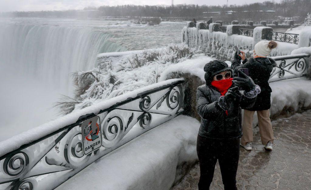 A woman with a red scarf over her face and a black coat on with the hood up takes a selfie from a platform overlooking Niagara Falls. The waterfall is frozen. There is a sign on the railings above the platform which says 'Danger'.