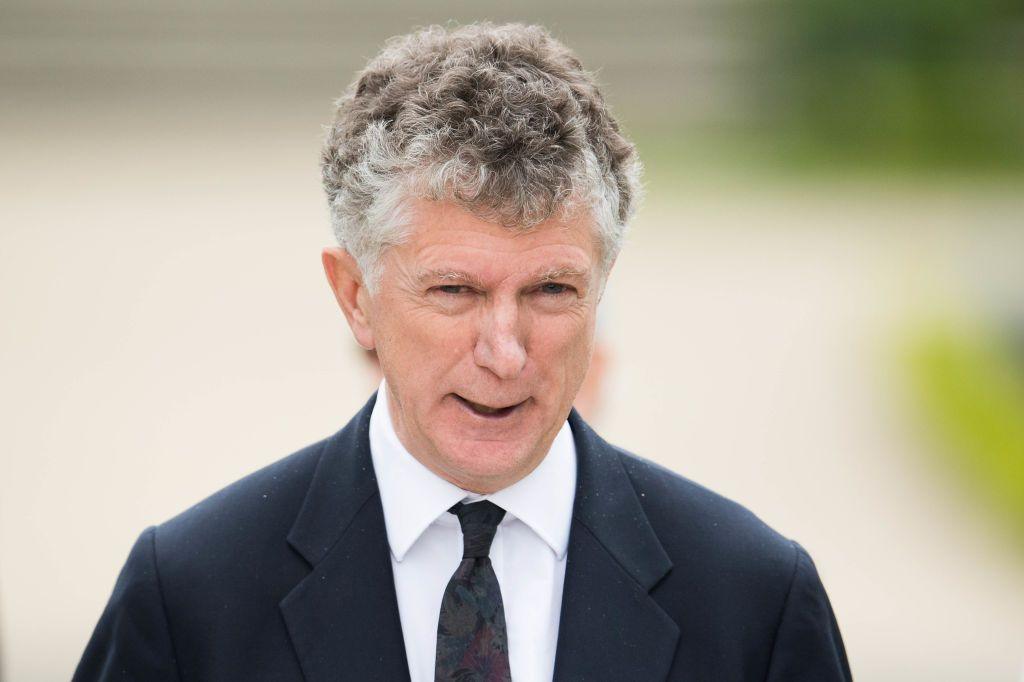 Mid-shot of Jonathan Powell with curly ash hair wearing a black suit, white shirt and black tie.