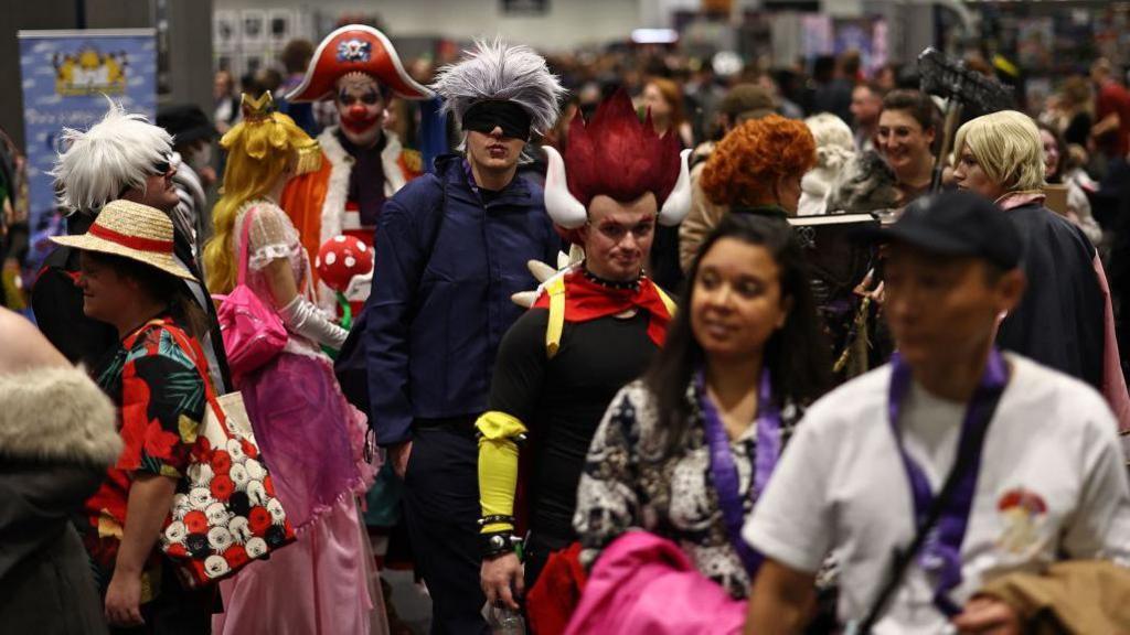 Groups of fans, many in colourful costumes, making their way across the floor of a convention 