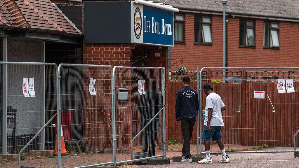 Two men walk through a fence into a hotel compound with a sign above reading 'The Bell Hotel'