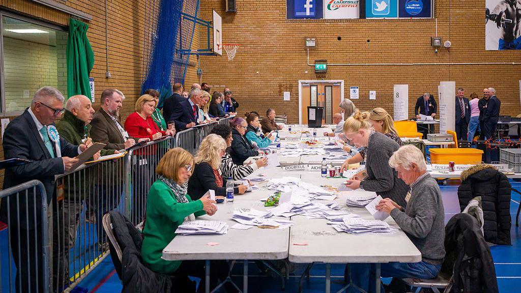 A general view of the count at Caerphilly Leisure Centre. There are people around a table counting papers while others look on.