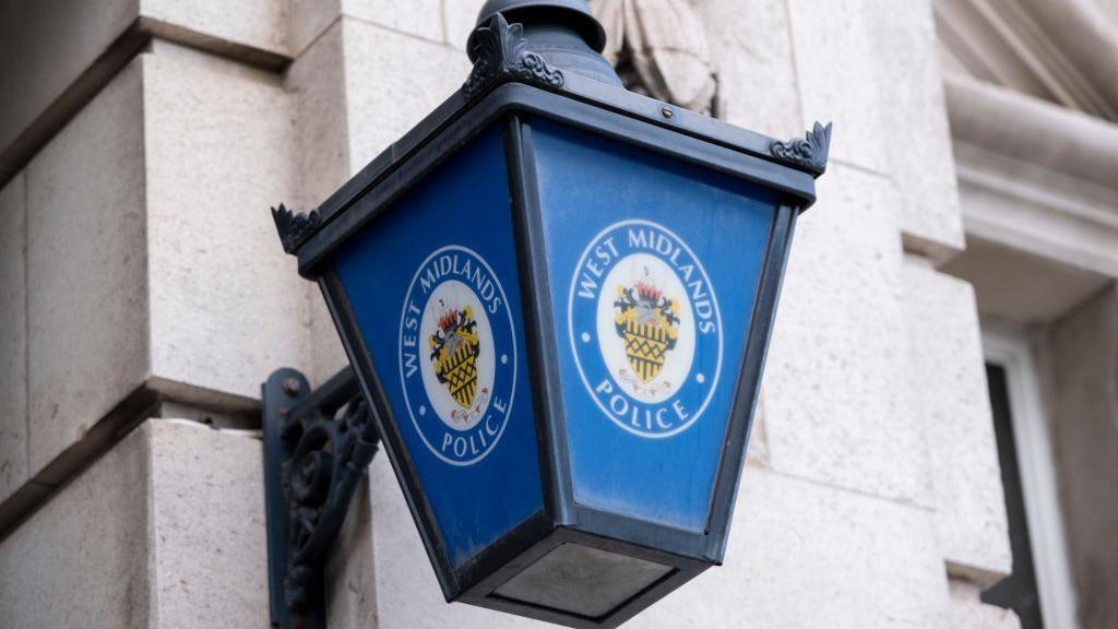 A blue lamp hanging on a grey stone wall. the lamp has black ironwork and the West Midlands police badge on it including the wording "West Midlands Police"