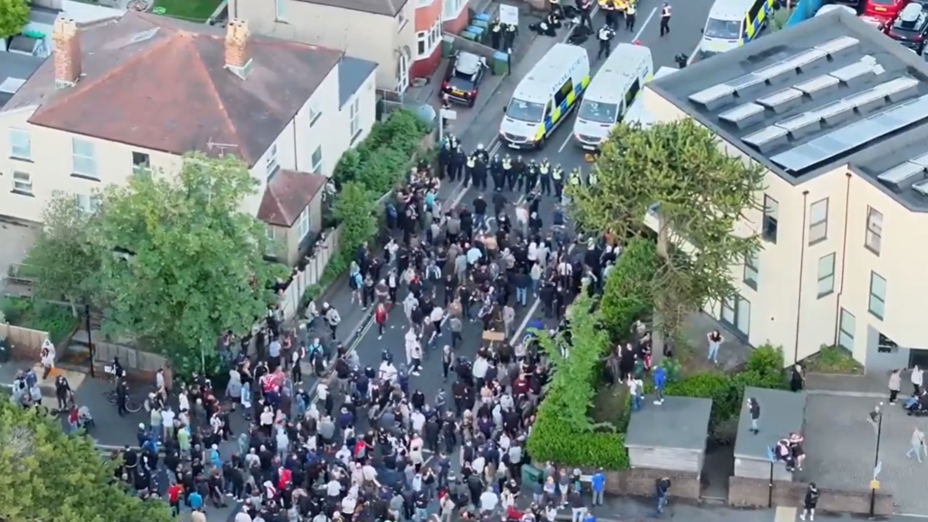 Aerial footage of a crowd facing a line of officers wearing riot gear. Behind them are several riot vans