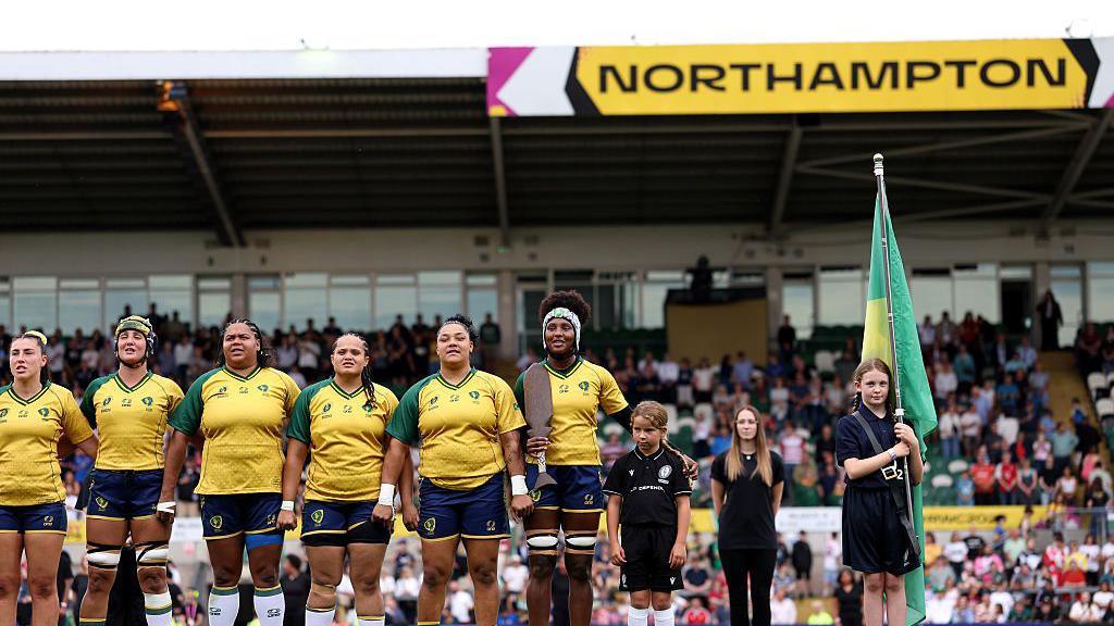 Brazilian rugby players stand in a line in yellow shirts and blue shorts. Next to them is a child mascot in black, while another child in black is holding a Brazil flag. Behind them in a stand full of fans and a sign that reads "Northampton".
