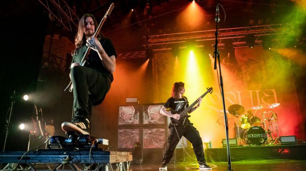 A guitarist stands at the front of a stage bathed in a mix of orange, red and green stage lights. He has one foot resting on a pedalboard which is slightly elevated above stage level. His long hair cascades down his shoulders as he plays notes. In the background, a bassist in a black band t-shirt stands side-on, playing along with his bandmate.