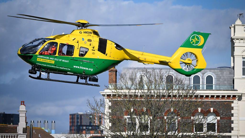 A yellow and green hampshire and isle of wight air ambulance helicopter hovering in the sky about building height above a city. There are crew members in orange onboard