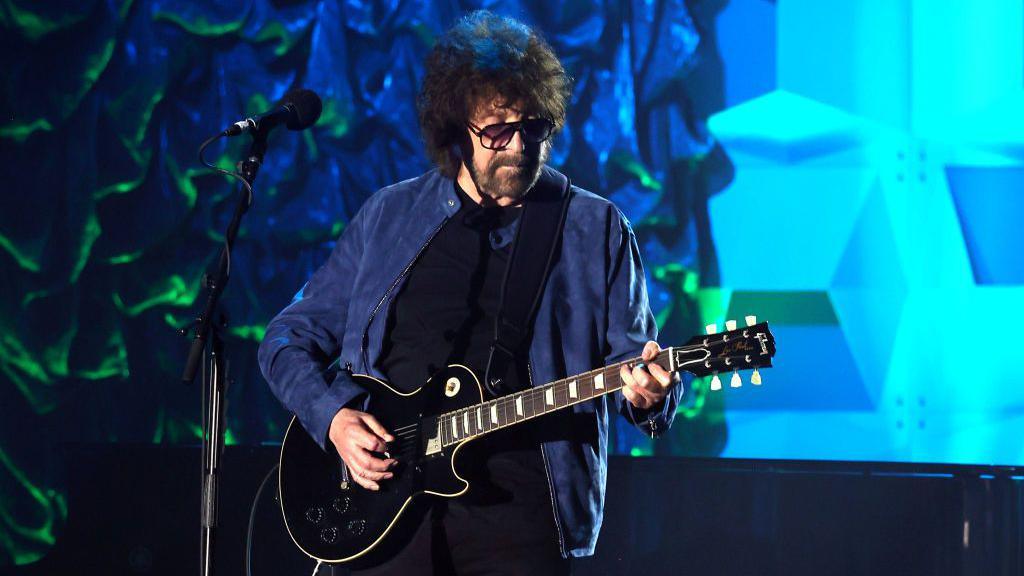 A man playing an electric guitar on a stage. The man has curly brown hair and a full beard. He is wearing large dark glasses and wearing a blue jacket. The stage has an illuminated blue and green backdrop.