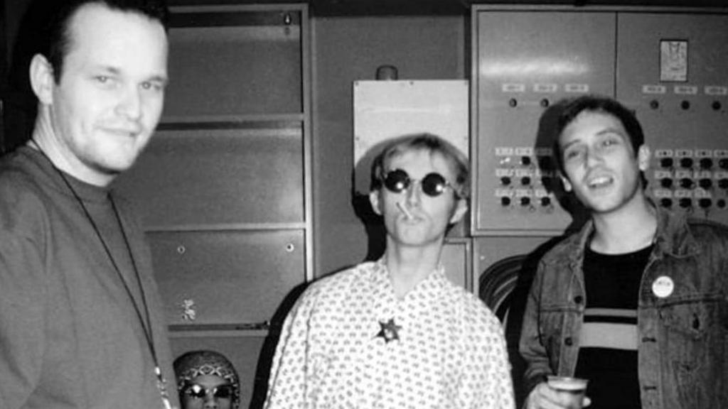 Black and white image of three men backstage at a concert. One man is smoking a cigarette and another has a beer in his hand