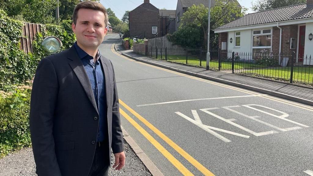 A man with short brown hair, wearing a dark jacket and blue shirt standing next to a road. 