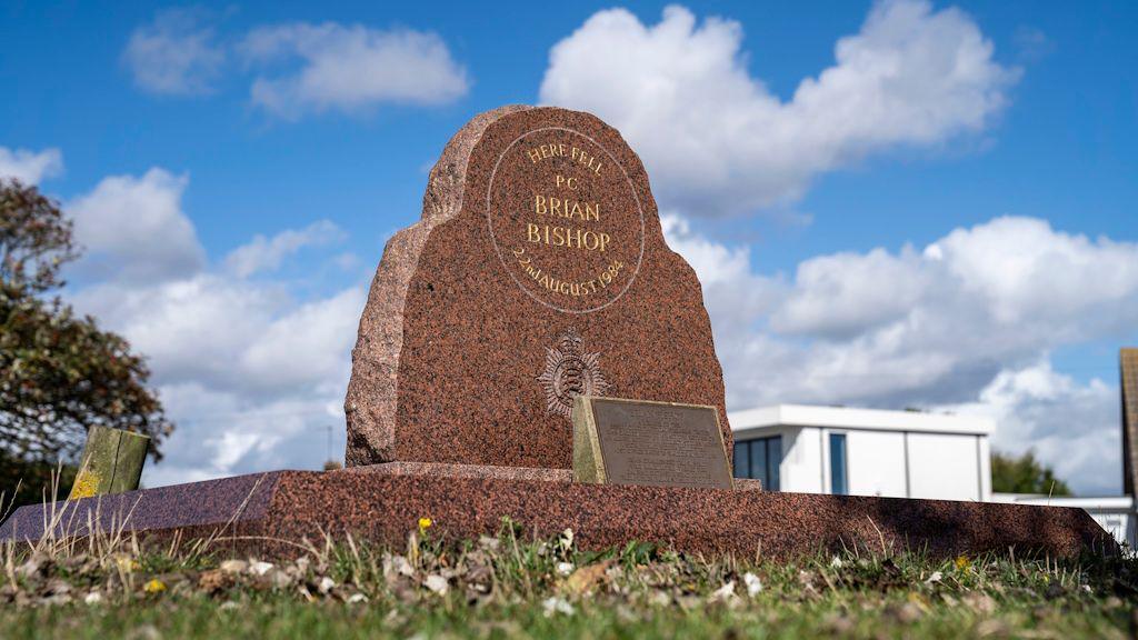 A memorial stone for PC Brian Bishop. It is made of red stone and bares his name and the date he passed away. A police building can be seen in the distance as well as a tree.