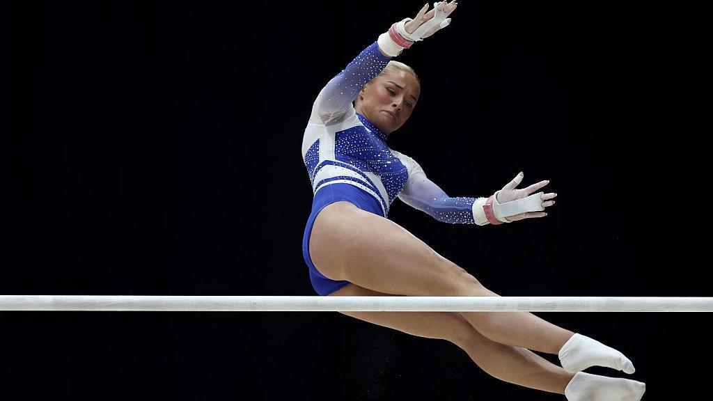 Ruby Evans in a blue and white leotard in the air as she competes on the uneven bars