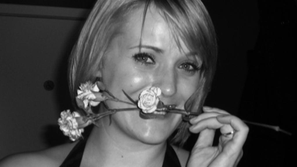 A black and white photo of a woman holding flowers between her teeth. She has blond hair in a short bob and is wearing a black halter neck top.