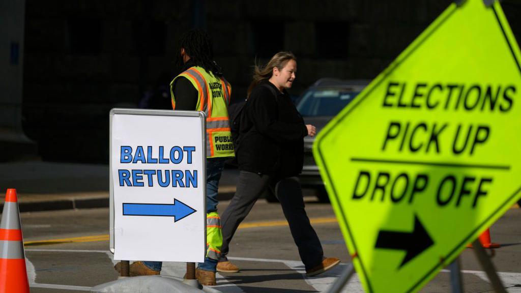 Woman walks past a sign saying 'Ballot return'