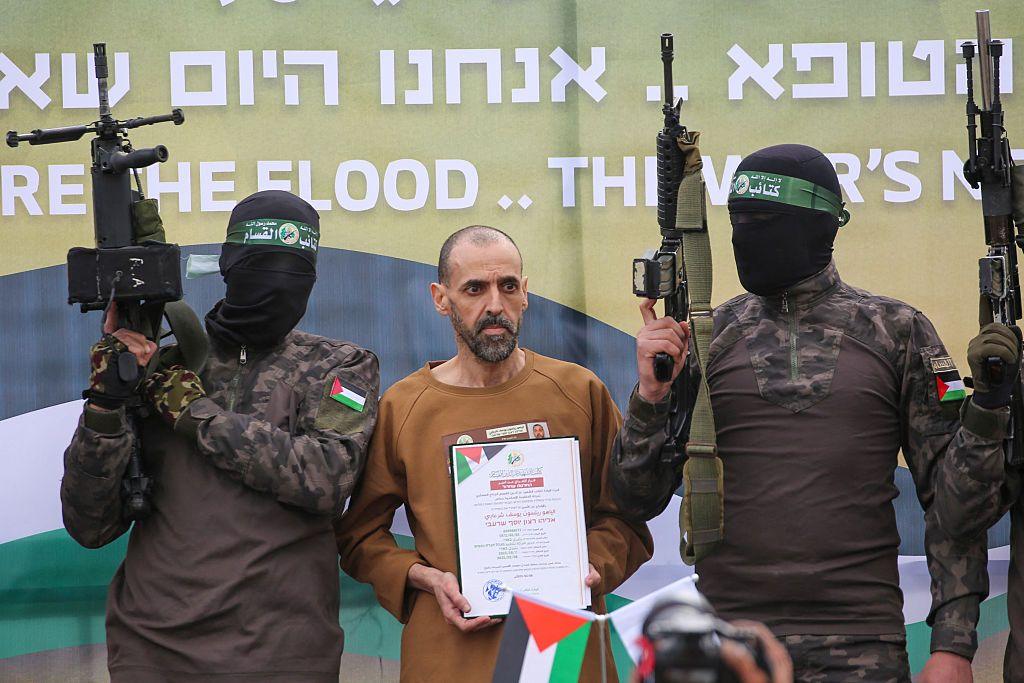 Heavily armed Hamas fighters escort a thin Israeli hostage Eli Sharabi on a stage before releasing him to Israel. The fighters have their faces masked, in military uniforms and are holding their guns in the air. Mr Sharabi is holding up a piece of paper and is wearing a brown shirt.