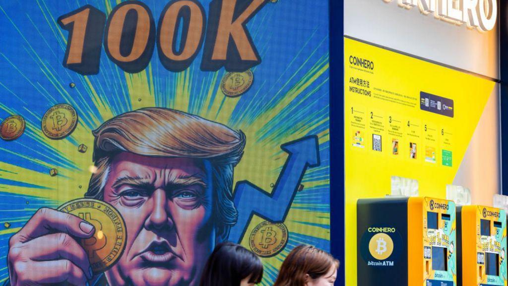 A colourful cartoon image of Donald Trump shown holding a Bitcoin token with "100k" displayed in big, bright lettering above it on a storefront in Hong Kong.