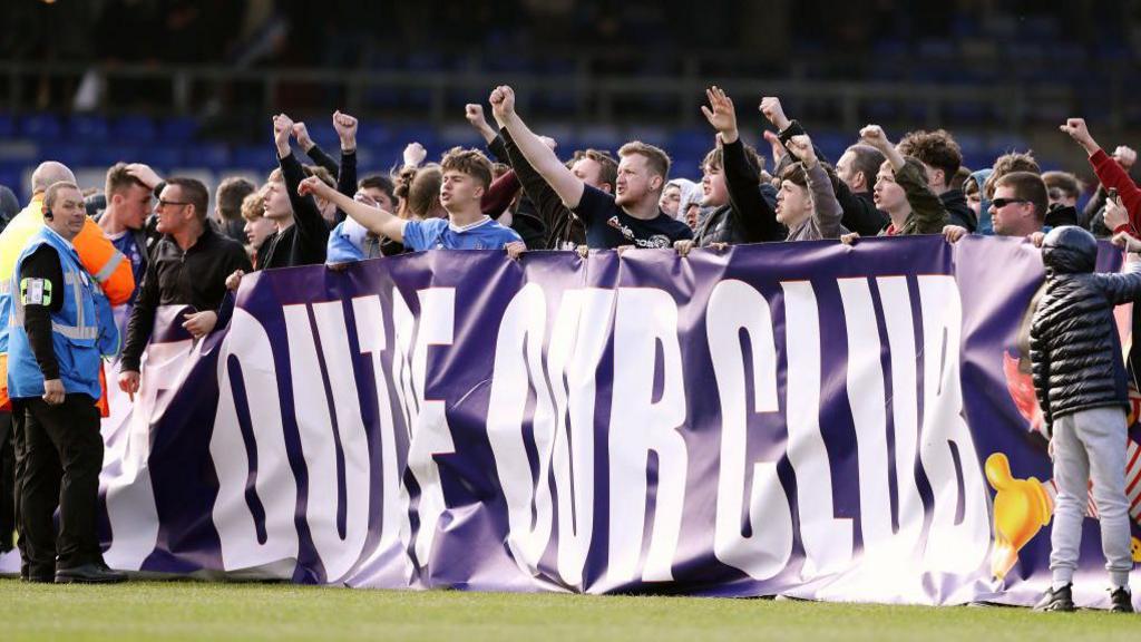 Oldham Athletic fans display a banner reading Get Out of Our Club, aimed at the club's then owner Abdallah Lemsagam, in an on-pitch protest at their home stadium Boundary Park
